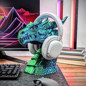 May include: A green and blue dragon head headphone stand. The dragon's head is facing to the left and has a white pair of headphones resting on its head.
