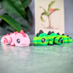 May include: Two articulated dinosaur toys. One is pink with red accents, the other is green and yellow with black accents. Both have large, cartoonish eyes and are lying on a gray surface. A botanical print is in the background.