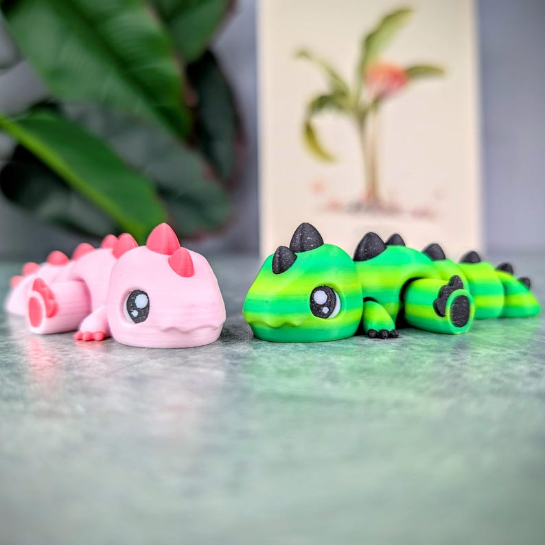 May include: Two articulated dinosaur toys. One is pink with red accents, the other is green and yellow with black accents. Both have large, cartoonish eyes and are lying on a gray surface. A botanical print is in the background.