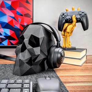 May include: A black skull-shaped headphone stand with a pair of black headphones resting on it. The stand is on a wooden desk with a keyboard, mouse, and a gold-colored decorative object in the background.