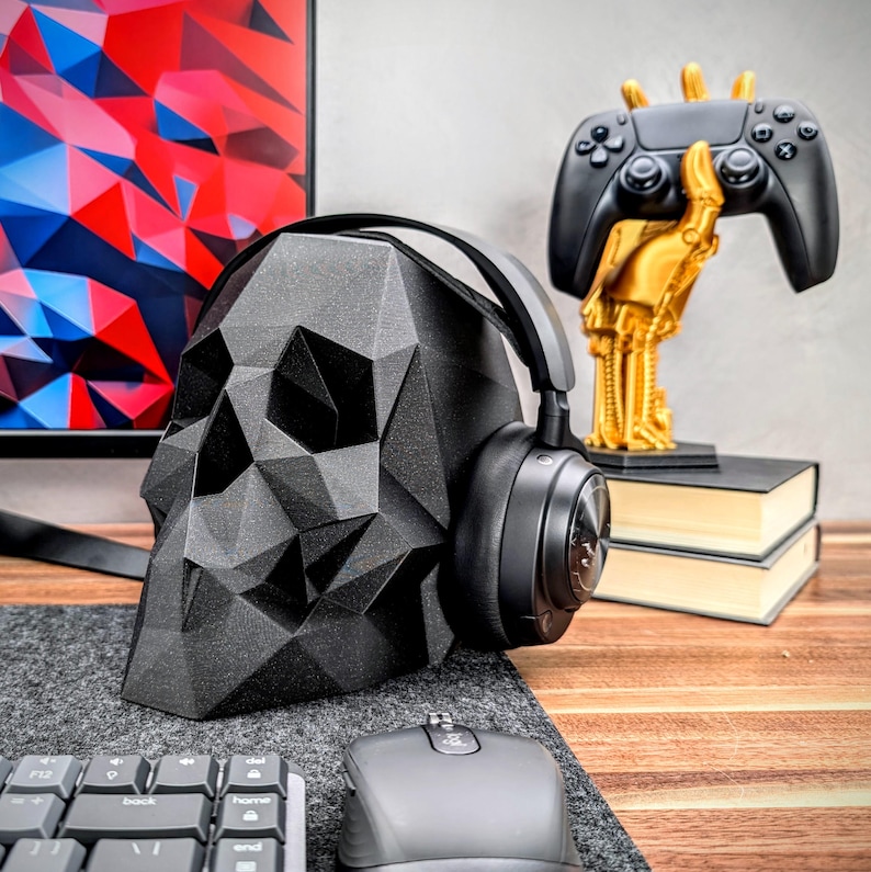 May include: A black skull-shaped headphone stand with a pair of black headphones resting on it. The stand is on a wooden desk with a keyboard, mouse, and a gold-colored decorative object in the background.