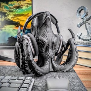 May include: A black octopus-shaped headphone stand with a pair of black over-ear headphones resting on its head. The stand is made of a 3D printed material and has a textured surface.