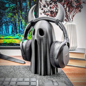 May include: A black ghost-shaped headphone stand with horns. The stand is made of 3D printed plastic and is designed to hold over-ear headphones.
