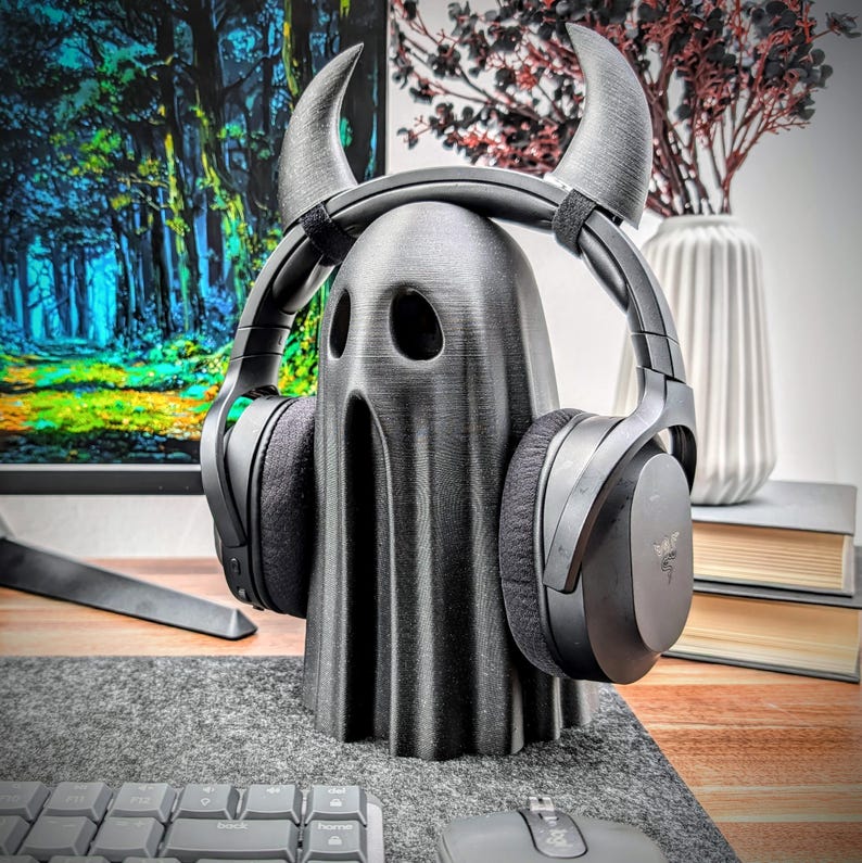 May include: A black ghost-shaped headphone stand with horns. The stand is made of 3D printed plastic and is designed to hold over-ear headphones.