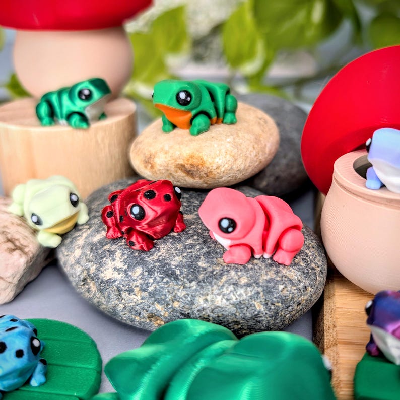 May include: A collection of colorful, miniature frog figurines in various shades of green, red, pink, and blue. The frogs are posed on small rocks and wooden platforms, with a mushroom-shaped decoration in the background. The frogs have large, black eyes.