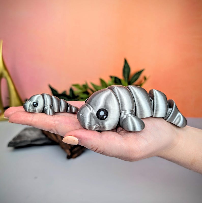 May include: Two articulated silver manatee toys with black eyes are displayed. The larger manatee is in the foreground, and the smaller one is behind it. The toys are made of segmented pieces, allowing them to bend and move. The background is a soft gradient of pink and orange.