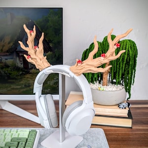 May include: A white headphone stand with a tree branch design, adorned with red mushroom accents. The stand holds a pair of white and gray headphones. The design is whimsical and decorative.