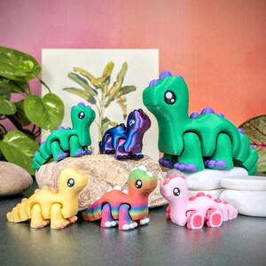 Op de afbeelding: Een verzameling kleurrijke, beweegbare dinosaurusfiguren. De dinosaurussen zijn in verschillende kleuren, waaronder groen, geel, roze en regenboog, met paarse accenten. Ze zijn geplaatst op kleine rotsen en een donkere ondergrond, met een botanische print op de achtergrond.