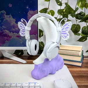 Pode incluir: Auscultadores brancos com detalhes de borboleta roxa, colocados num suporte branco em forma de lua crescente com base de nuvem roxa. O suporte est&aacute; sobre uma mesa branca com um teclado e um rato.