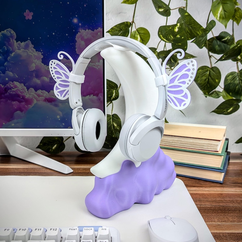 Pode incluir: Auscultadores brancos com detalhes de borboleta roxa, colocados num suporte branco em forma de lua crescente com base de nuvem roxa. O suporte est&aacute; sobre uma mesa branca com um teclado e um rato.