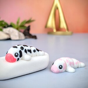 May include: Two handcrafted polymer clay sculptures of worm-like figures. One is white with black spots and a pink nose, lying on a white base. The other is white with pink spots and a pink nose. The background is a blurred pink and orange gradient.