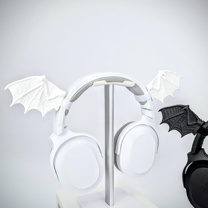 May include: White wireless headphones with attached white bat wings are displayed on a silver stand. A black headphone with black bat wings is also visible. The headphones have a padded headband and earcups.