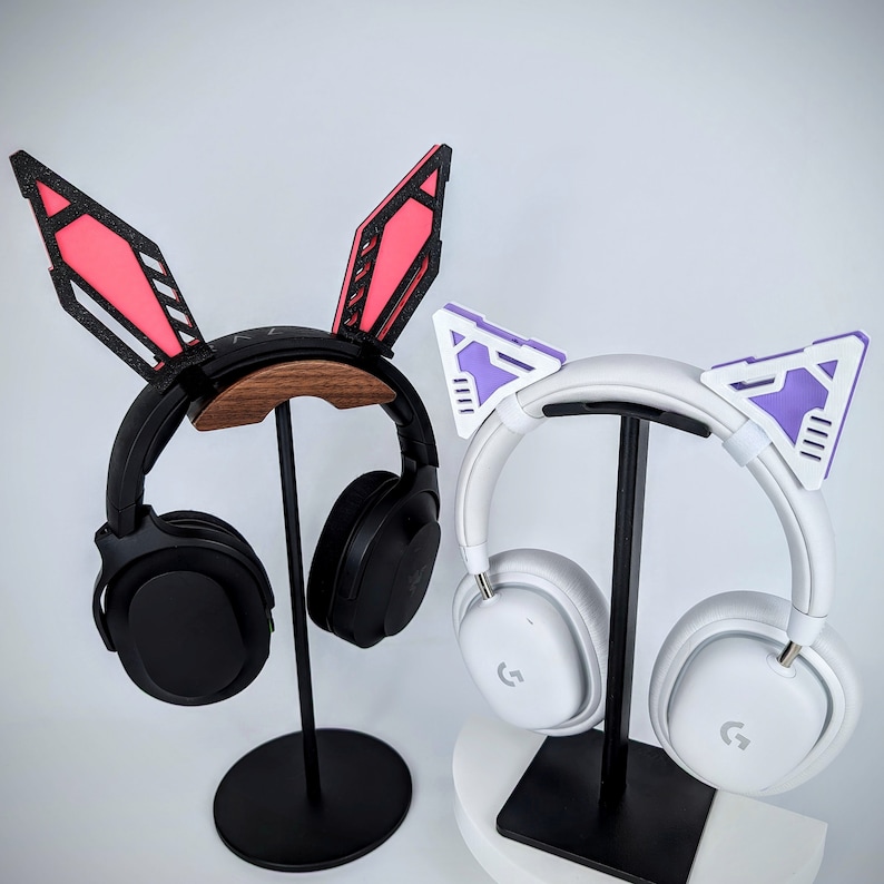 Pode incluir: Dois conjuntos de fones de ouvido com acess&oacute;rios decorativos para as orelhas. Um conjunto apresenta fones de ouvido pretos com detalhes de orelhas de coelho rosa e preto. O outro tem fones de ouvido brancos com detalhes de orelhas de gato roxo e branco. Ambos est&atilde;o em suportes pretos.