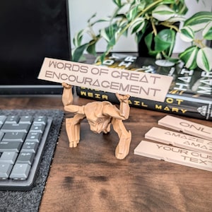 May include: A light brown 3D-printed figurine of a stylized humanoid figure with arms raised, holding a rectangular sign that reads "WORDS OF GREAT ENCOURAGEMENT" in dark brown lettering. Two additional blank sign inserts are visible nearby.