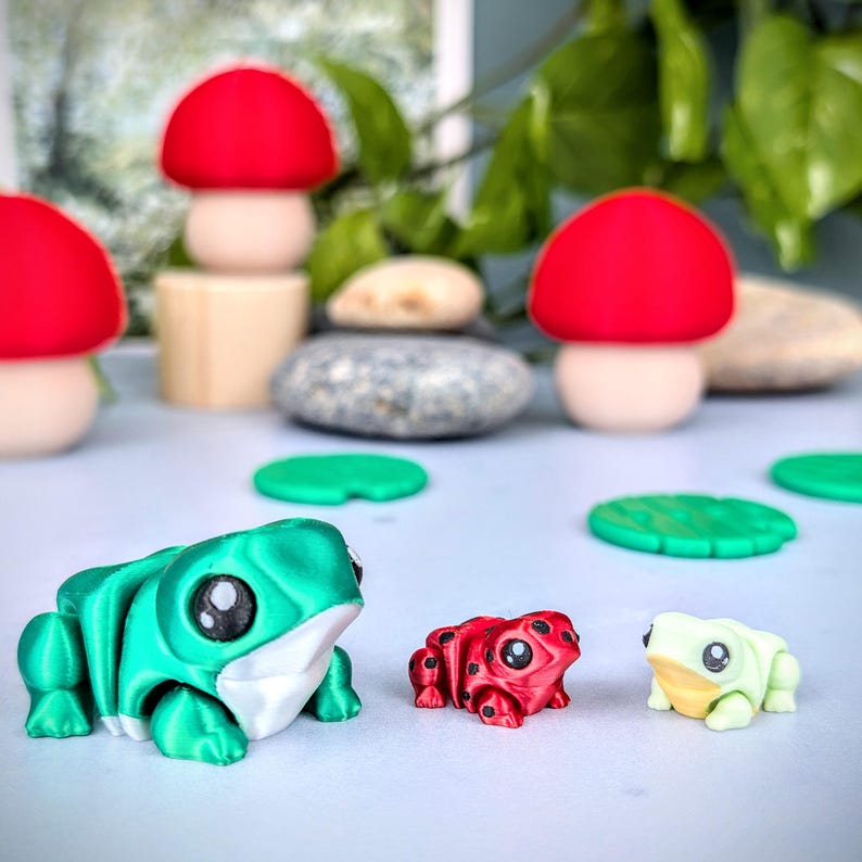 May include: A collection of whimsical, colorful figurines. The image features a large, green frog, a small red frog, and a small, light green frog. Also present are red-capped mushroom figurines and green lily pads. The background includes stones and greenery.