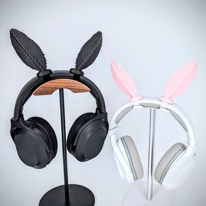 May include: Two headphone stands with bunny ear shaped holders. The stand on the left has a black headphone with a wooden base. The stand on the right has a white headphone with a silver base.
