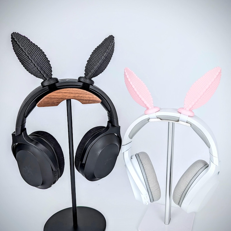 May include: Two headphone stands with different colored bunny ear headphone holders. The stand on the left has a black headphone with a wooden base and black bunny ears. The stand on the right has a white headphone with a silver stand and pink bunny ears.