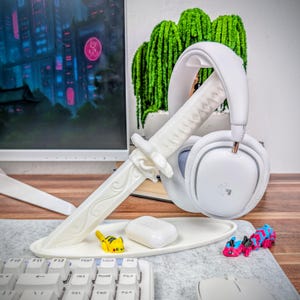 May include: A white headset rests on a decorative white katana-shaped stand. A small white tray holds a white charging case and a yellow and black toy. A keyboard and monitor are in the background.
