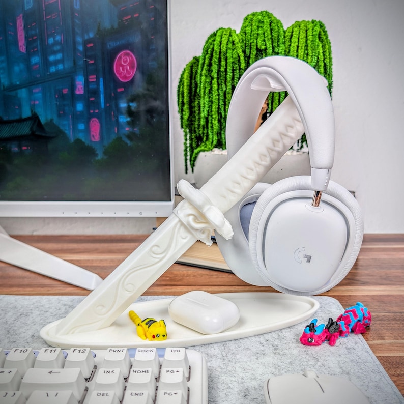 May include: A white headset rests on a decorative white katana-shaped stand. A small white tray holds a white charging case and a yellow and black toy. A keyboard and monitor are in the background.