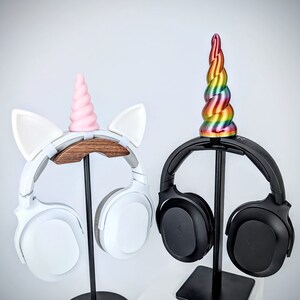 May include: Two headphone stands with unicorn-themed toppers. One stand is black with a rainbow unicorn horn and the other is white with a pink unicorn horn. Both stands have headphones attached.