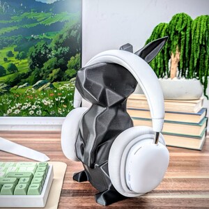 May include: A black, geometric rabbit-shaped headphone stand with white over-ear headphones resting on it. The stand is on a wooden surface.