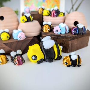 May include: A collection of hand-painted bee figurines in various colors, including yellow, black, blue, and iridescent. The figurines are arranged on a wooden surface and some are in a beehive-shaped container.
