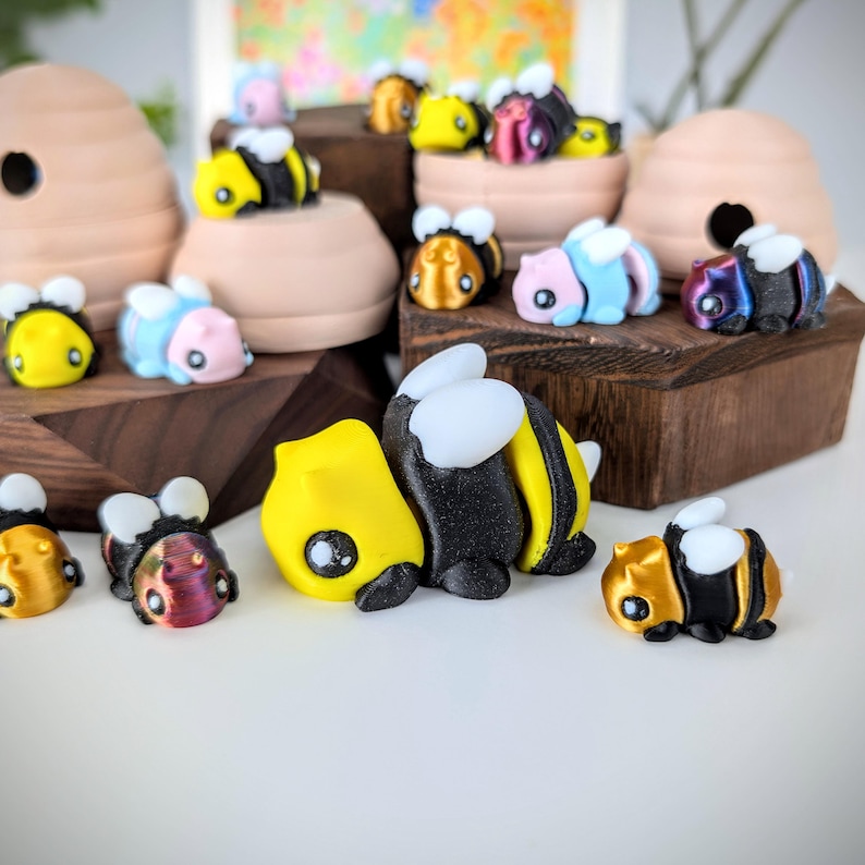 May include: A collection of hand-painted bee figurines in various colors, including yellow, black, blue, and iridescent. The figurines are arranged on a wooden surface and some are in a beehive-shaped container.
