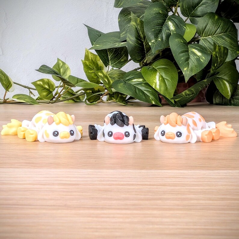 May include: Three small, hand-painted ceramic figurines of cows. One cow is black and white, one is yellow and white, and one is orange and white. The cows are lying down with their heads facing forward.