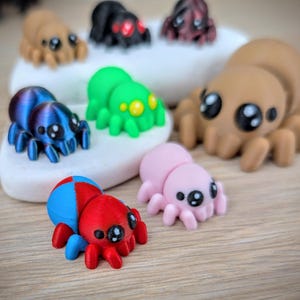 May include: A collection of colorful, cartoonish spider figurines. The spiders are in various colors, including blue, green, pink, brown, red, and black. Each spider has large, expressive eyes and a cute, friendly appearance. The figurines are displayed on a white stone and a wooden surface.