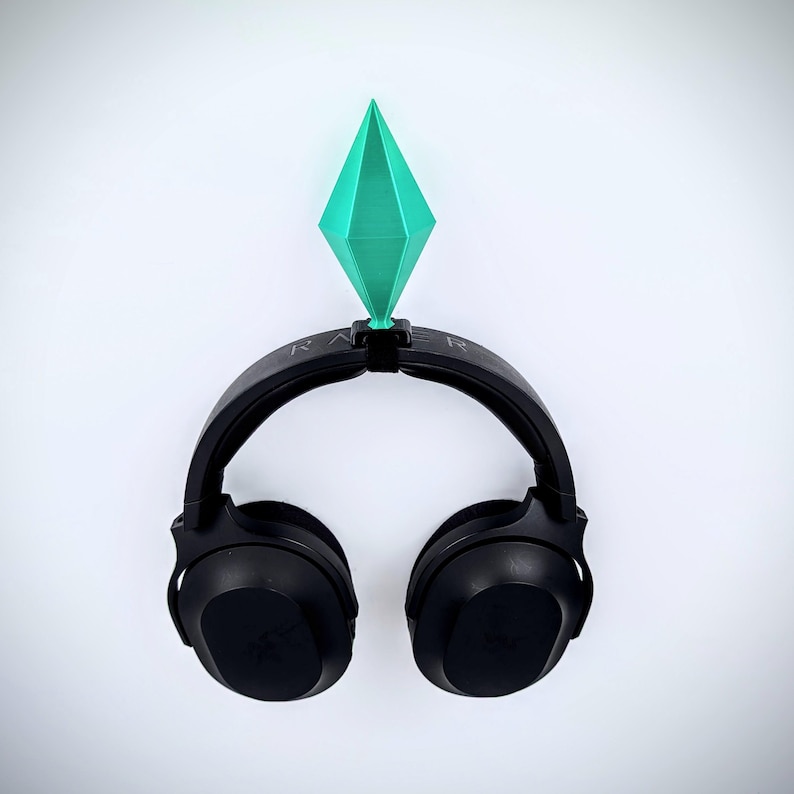 May include: Black headphones with a green diamond-shaped object on top. The headphones are hanging on a white wall.