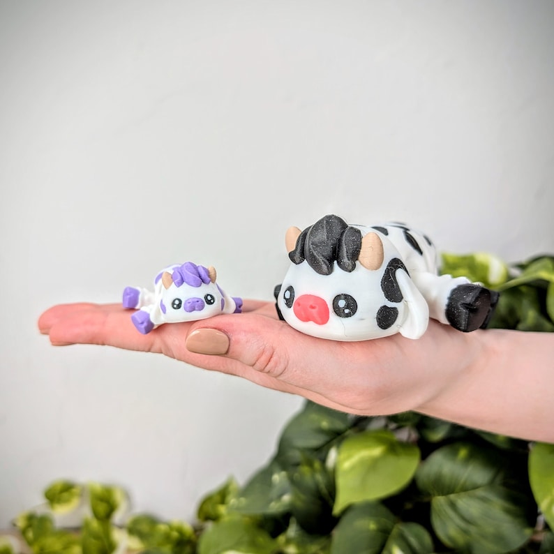 May include: Two small, hand-painted cow figurines. The larger cow is black and white with a pink nose and black eyes. The smaller cow is white with purple accents and black eyes.