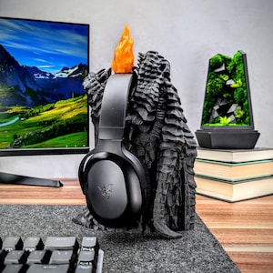 May include: Black gaming headset with a black dragon-shaped stand. The stand has a flame-shaped orange topper. The headset is resting on the stand.