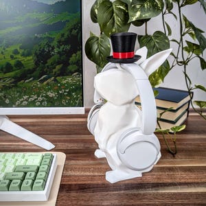 May include: White rabbit-shaped headphone stand with a black top hat and red band. The stand is holding a pair of white headphones.