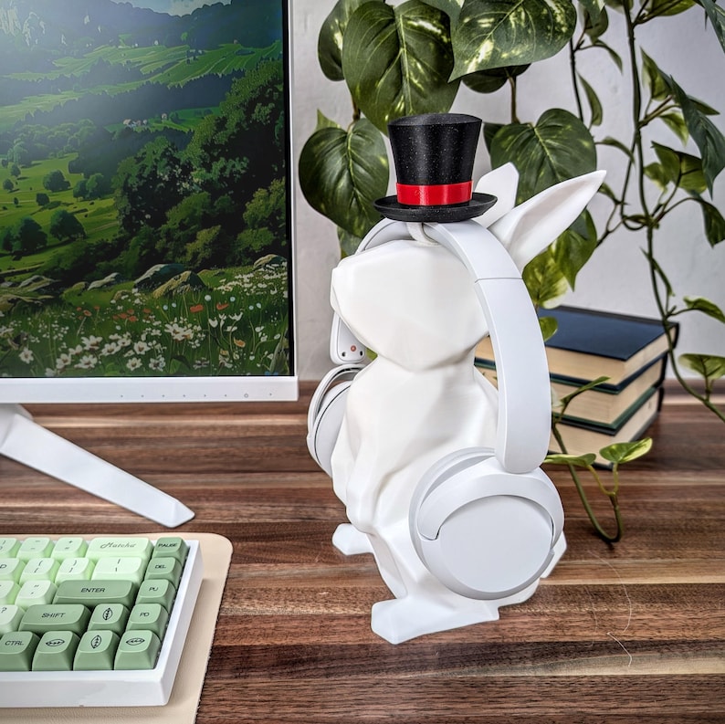 May include: White rabbit-shaped headphone stand with a black top hat and red band. The stand is holding a pair of white headphones.