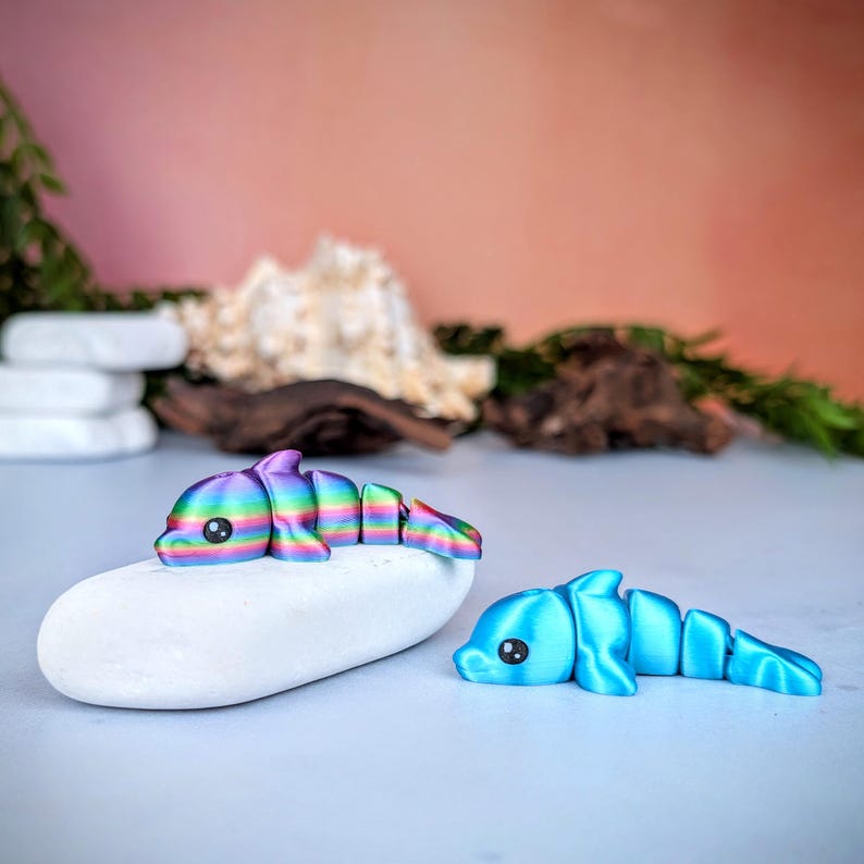 May include: Two articulated dolphin figurines. One is resting on a white stone and is a rainbow of colors. The other is a solid light blue. Both have black eyes and are segmented to allow for movement. The background is a soft peach color.