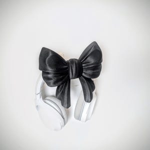 May include: A pair of white wireless headphones with gray ear pads, accented by a large, black satin bow. The headphones are positioned on a white background, creating a clean and modern aesthetic. The bow adds a touch of elegance.