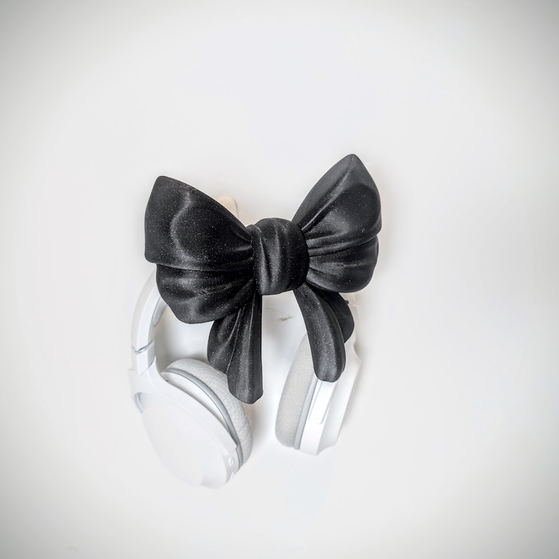 May include: A pair of white wireless headphones with gray ear pads, accented by a large, black satin bow. The headphones are positioned on a white background, creating a clean and modern aesthetic. The bow adds a touch of elegance.