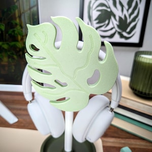 May include: A white headphone stand with a green monstera leaf design. The stand has two white headphones hanging from it.