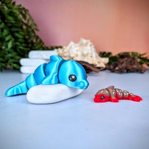 May include: A blue 3D-printed dolphin toy resting on a white stone. The dolphin has segmented body parts and black eyes. A smaller red and brown segmented toy is next to the dolphin. The background is a soft pink.
