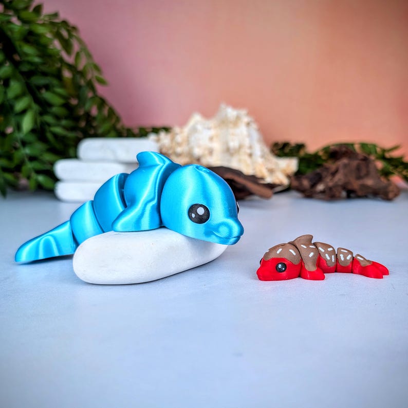 May include: A blue 3D-printed dolphin toy resting on a white stone. The dolphin has segmented body parts and black eyes. A smaller red and brown segmented toy is next to the dolphin. The background is a soft pink.