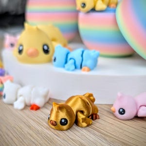 May include: A collection of colorful, small animal figurines, including yellow ducklings, blue and white creatures, and a gold-colored cat-like figure. The figurines are arranged on a light-colored surface, with pastel-colored egg-shaped decorations in the background.