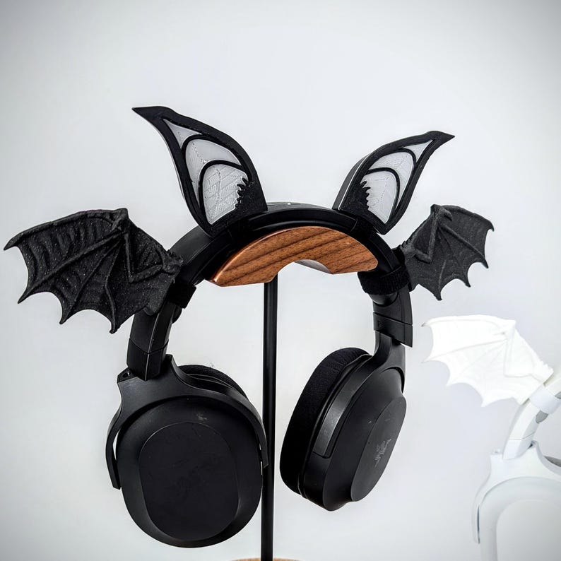 May include: Black headphones with bat wing and ear accents rest on a black headphone stand with a wooden accent. The bat wings are black with textured details. The ear accents are black with white detailing.