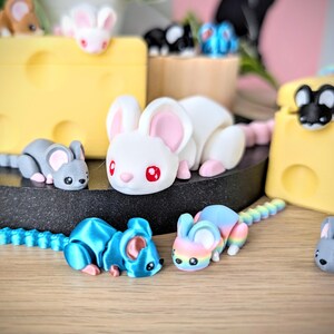 May include: A collection of small, colorful, hand-painted mouse figurines. The mice are made of a smooth, white, gray, blue, and rainbow colored material. The mice are arranged on a black surface with a yellow and white background.