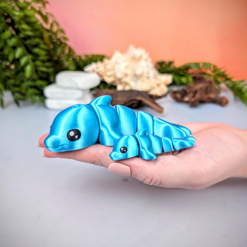 May include: A pair of articulated, light blue dolphin figurines with black eyes. The larger dolphin is positioned above a smaller one, both held in a hand. The background features greenery and decorative elements.
