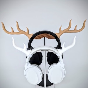 May include: A white headset with a black headband is displayed on a wooden stand with two antlers. The stand is made of wood and has a brown finish.