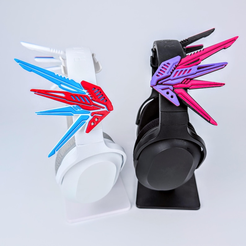 May include: Two gaming headsets, one white and one black, each adorned with decorative wing-shaped attachments. The white headset features blue and red accents, while the black headset has pink and purple details. Both are displayed on white stands.