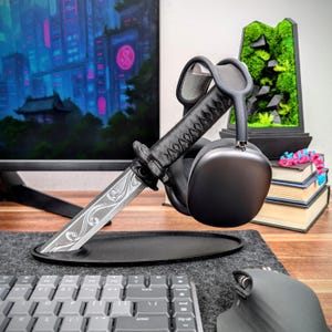 May include: A black headphone stand shaped like a katana sword. The stand holds a pair of black headphones. The sword blade has an engraved design. The stand sits on a black oval base on a gray desk mat.
