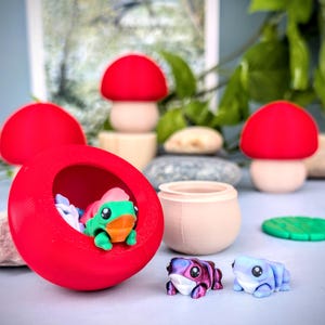 May include: A whimsical collection of miniature figurines and decor. A large red pod houses a green and orange frog figurine. Other items include mushroom-shaped decor, small frog figurines, and a small pot.