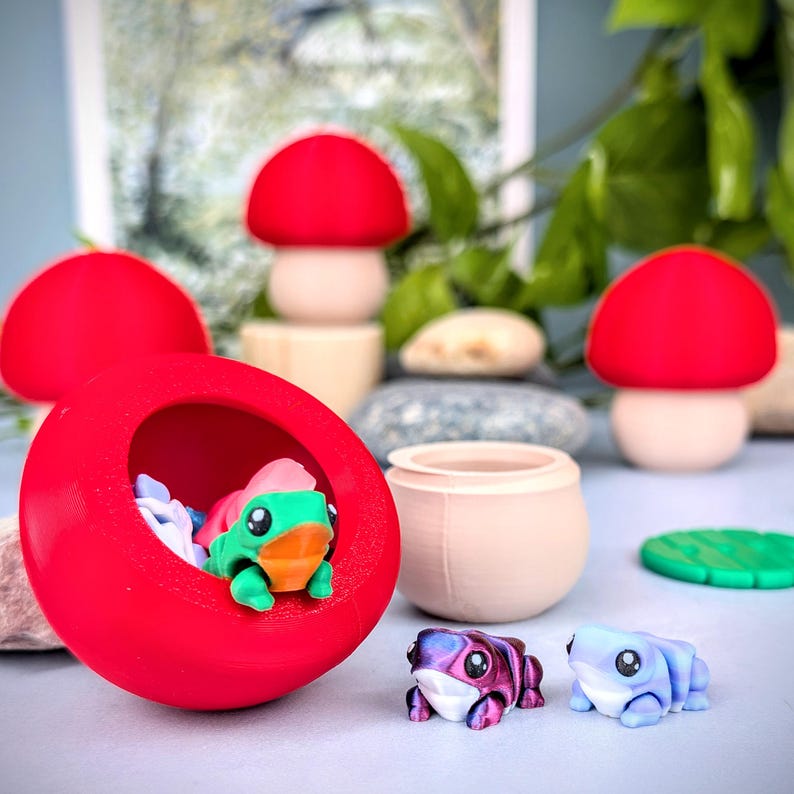 May include: A whimsical collection of miniature figurines and decor. A large red pod houses a green and orange frog figurine. Other items include mushroom-shaped decor, small frog figurines, and a small pot.