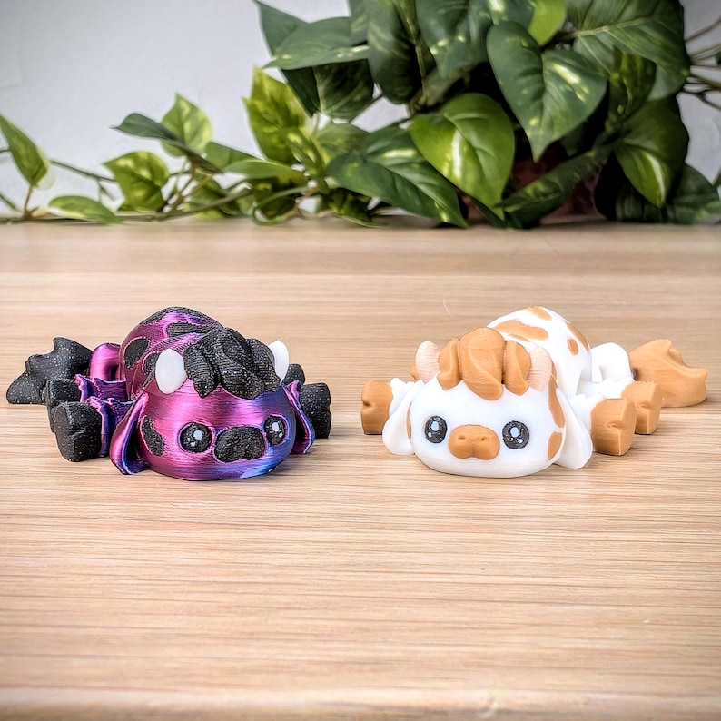 May include: Two small, hand-painted figurines. One is a purple and black creature with large eyes and horns. The other is a white creature with brown spots and large eyes. Both are lying on a wooden surface.